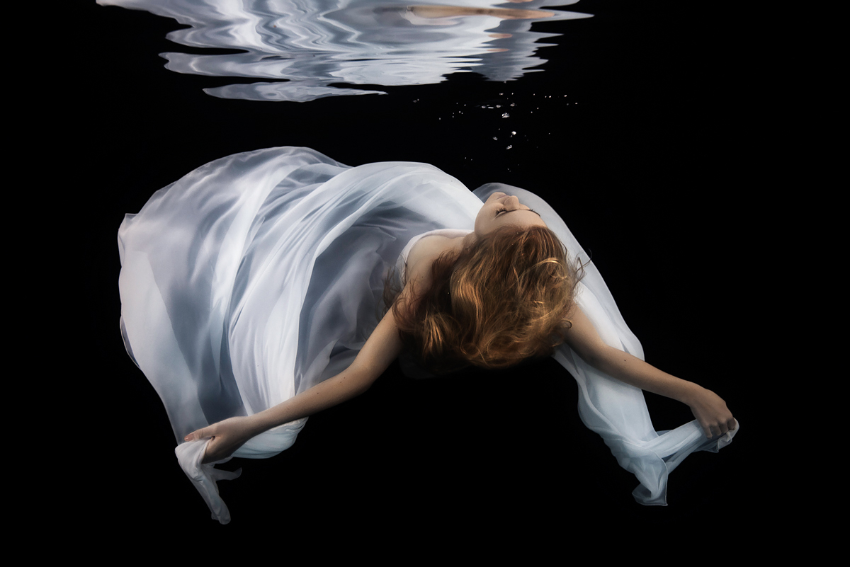 Underwater portrait of a beautiful bride in white gown sinking back down through the water, arms outstretched, black background, trash the dress shoot