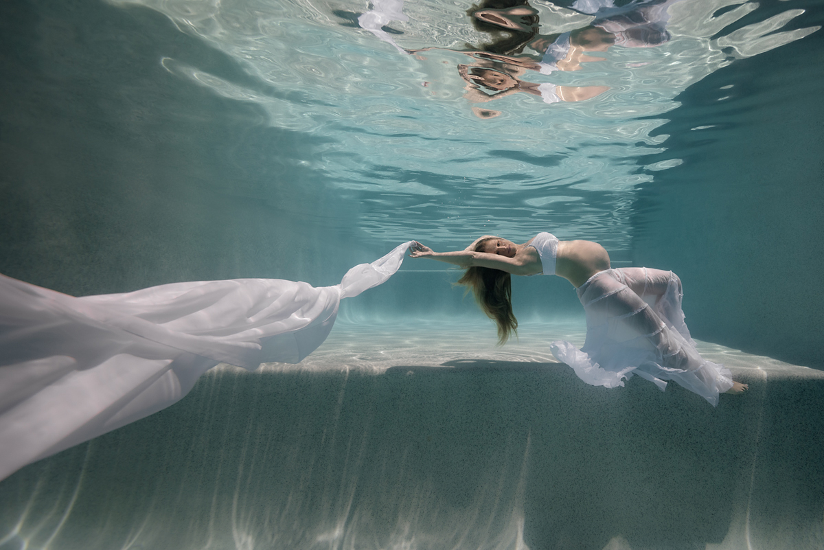 Underwater maternity portrait of a pregnant lady in a large swimming pool, wearing flowing white skirt and draping a long white chiffon scarf.