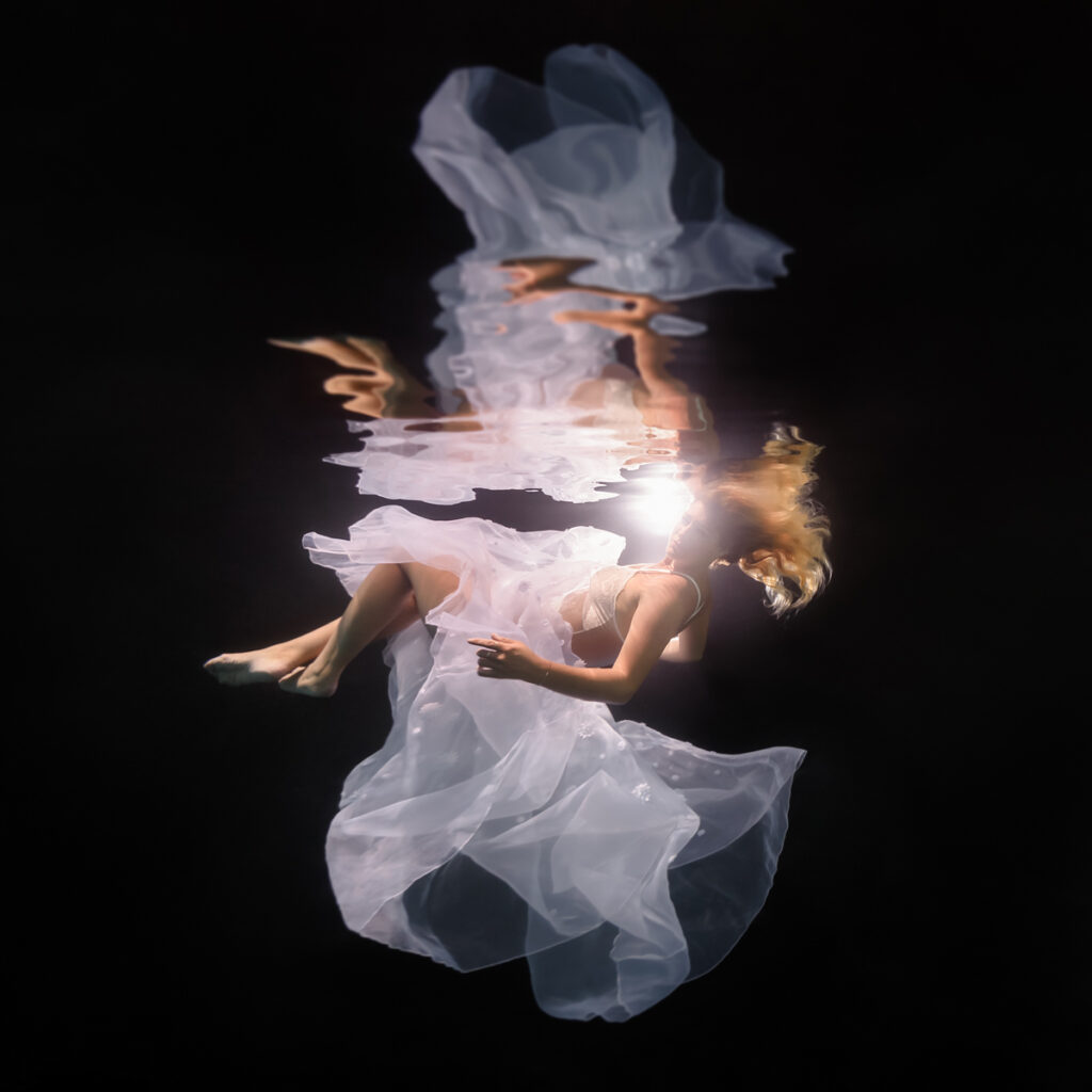 Woman underwater, blonde hair, floaty white chiffon dress, black background, backlit, with watery reflections