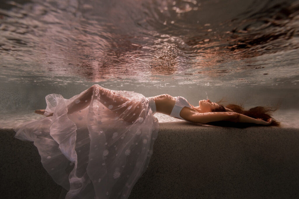 Beautiful woman wearing white sheer fabrics, laying on a ledge underwater in a swimming pool