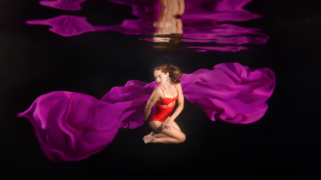 Colourful artistic underwater portrait of a woman twirling and twisting underwater, wearing an orange bodysuit, and surrounded by purple floating chiffon fabric; black background; reflections; photographed in Noosa, on the Sunshine Coast, Queensland, Australia