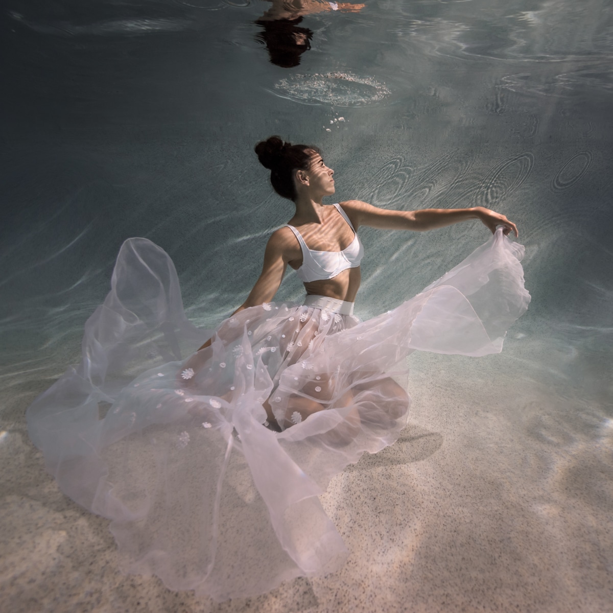 Underwater portrait photo of woman sitting at the bottom, wearing a floating sheer white skirt, with sunbeams