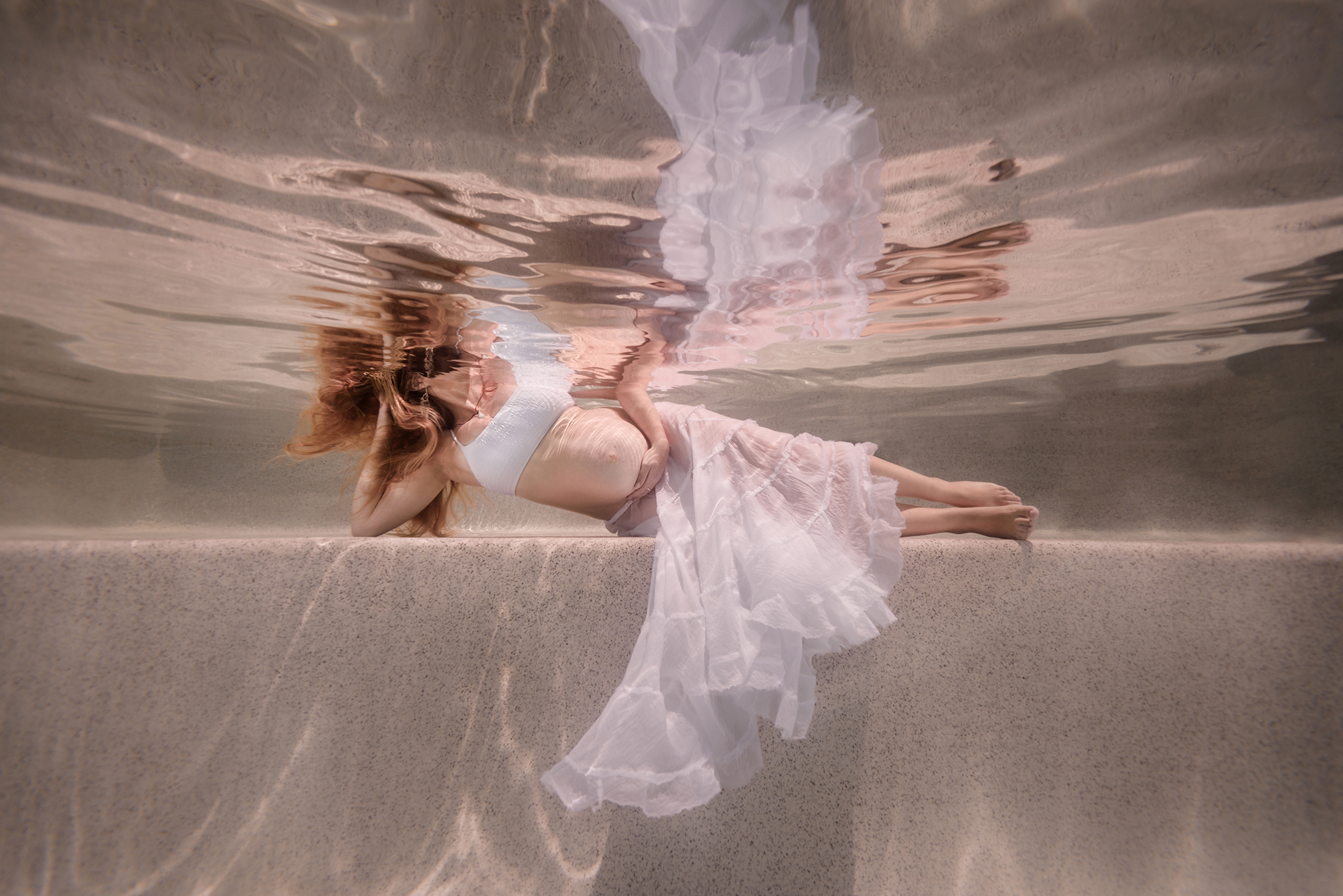 pregnant woman laying on her side underwater in a pool, wearing a white top and a white skirt