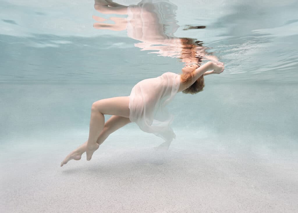pregnancy photo of a blonde woman floating peacefully underwater, draped in a white cloth