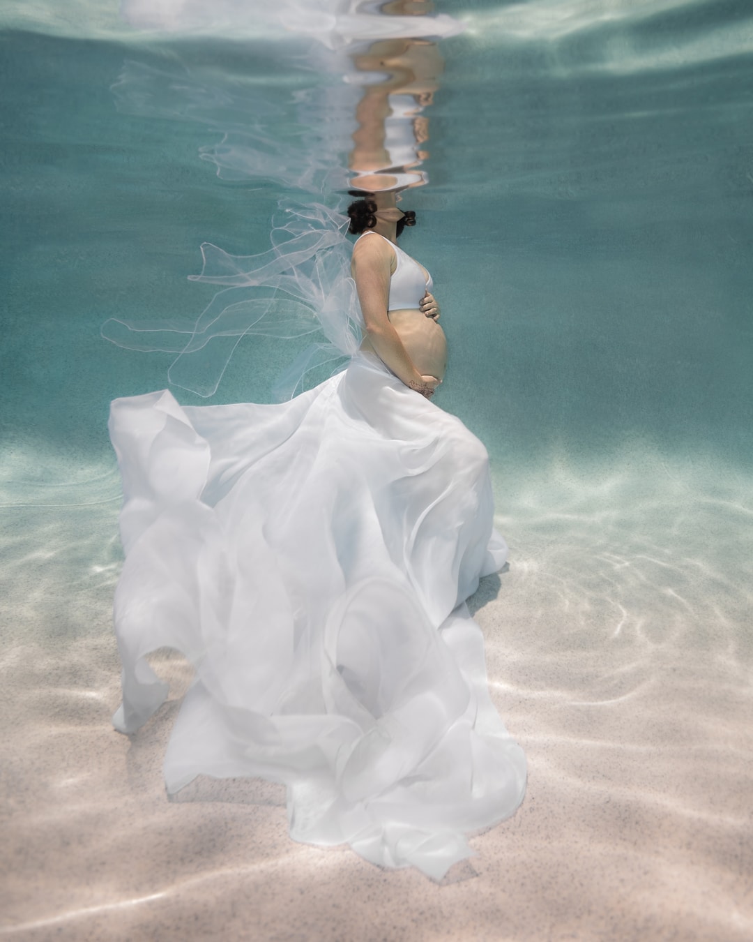 Underwater pregnancy portrait of a woman holding her belly and wearing a flowing white gown