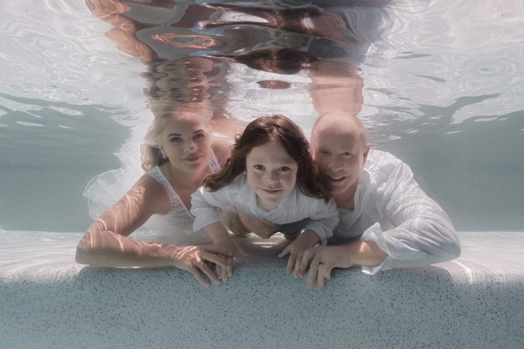 Underwater family portrait of a mother, father, and young son with their arms around each other, floating together in a soft, ethereal pose.